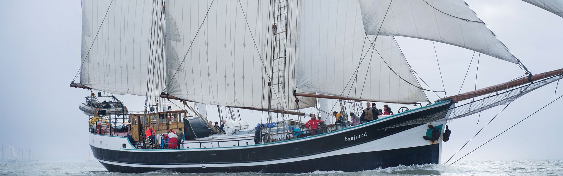 Der schwarz-weiße Rumpf des Segelschiffs Banjaard gleitet mit gesetzten Segeln durch das Waddenmeer. An Deck stehen Crew und Gäste bereit, um die Tau zu bedienen für das nächste Manöver.