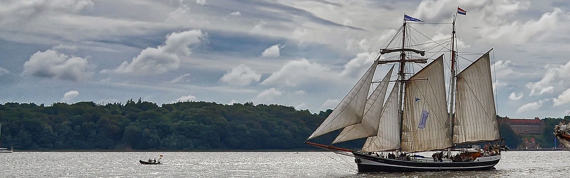 Die Banjaard segelt vor einer dichtbewaldten Steilküste. Die Wolken und Flaggen lassen einen windigen Tag erahnen, aber das Wasser ist unter dem Schutz der Küste ruhig.