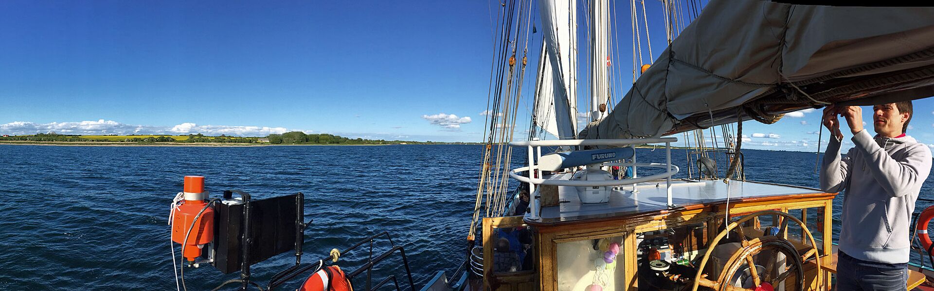 Inselpanorama Daenemark gesehen vom Achterdeck des Schiffes Banjaard: die dunkelblaue Ostsee vor dem Blau des Himmels und der grünen Vegetation der Insel.