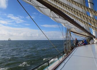 Blick unter dem gesetzten Schonersegel und den Tauen der Schot durch auf den Horizont und davor das dunkelblaue Meer mit weißen Schaumkronen.