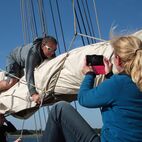 Ein Mann balanciert bäuchlings auf der Großgaffel und zieht die Persenning über das gefaltete Großsegel darunter um es einzupacken. Eine Frau macht ein Foto von ihm dabei.