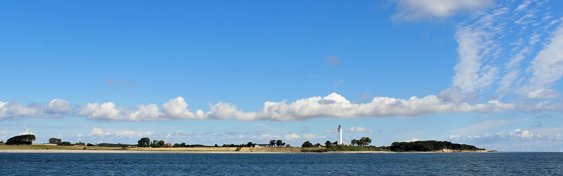 Blick vom Wasser auf ein stück dänischer Küste mit einem hohen, allein stehenden, weißen Leuchtturm umrahmt von gelben Getreidefeldern, grünen Bäumen und einenm hellblauen Himmel mit ein paar weißen Wolken.