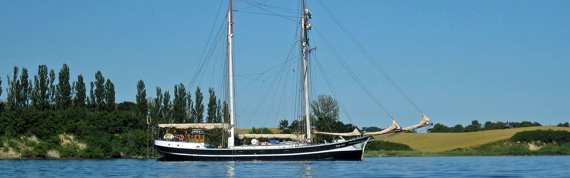 Vor der Küste mit grünen Bäumen und ockerfarbenen Getreidefeldern liegt an einem wolkenlosen Tag das Segelschiff Banjaard vor Anker. Alles wirkt ruhig, friedlich und entspannt.