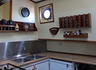 Schiffsküche auf der Banjaard mit einer Edelstahlspüle, weißen Unterschränken, Gefrierschrank und an den Wänden Regale und Halterungen für Vorratsbehältern, Kaffeefilter, Gewürze und Tassen, eine Wanduhr und ein Bullauge