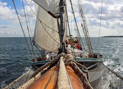 Blick vom Klüverbaum aus auf das Schiff Banjaard, das mit gesetzten Segeln unter einem teilweise bewölkten Himmel über ruhige Gewässer gleitet.