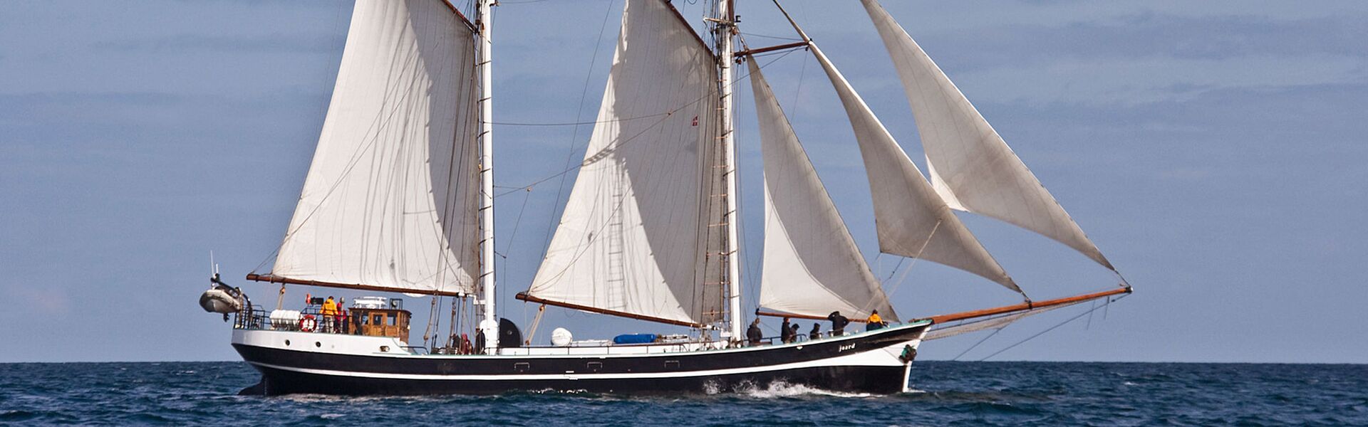 Das Segelschiff Banjaard unter Segeln auf der Ostsee: die Sonne beleuchtet die Segeln, an Deck stehen mehrere Gäste, die aufmerksam die Segel betrachten.