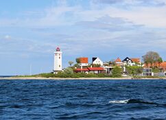 Malerische Aussicht auf einen weißen Leuchtturm auf einem Küstenvorsprung mit Strand und bunten Häusern, umgeben von tiefblauem Wasser und klarem Himmel.
