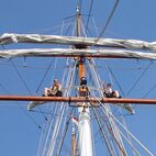 Zwei Personen sitzen auf der Saling eines Segelschiffs und winken vor einem klaren blauen Himmel, umgeben von Segeln und Takelage.