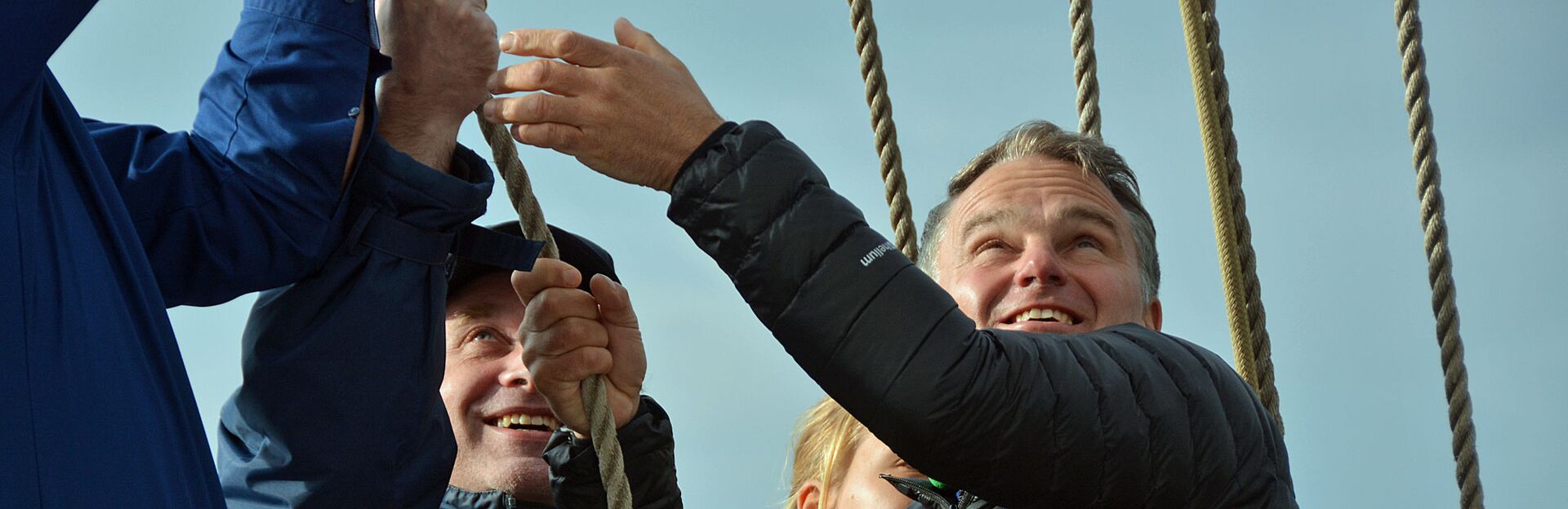 Drei Menschen in Jacken lächeln, während sie gemeinsam an einem Seil ziehen und in den klaren Himmel blicken. Die Stimmung ist fröhlich und kooperativ und lässt auf Teamarbeit schließen.