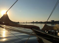 Sonnenaufgang über dem Fluss Warnow: im Vordergrund der Bug eines Segelschiffes und im Hintergrund die Skyline von Rostock