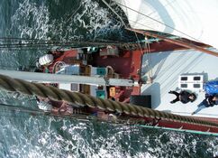 Luftaufnahme vom Mast aus vom Schiffsdecks der Banjaard mit Seilen und Segeln. Gäste liegen entspannt auf dem Deck und schauen zum Fotografen hoch. Um das Schiff herum ist das dunkelgrün erscheinende Meer von weißen Schaum gekrönt.