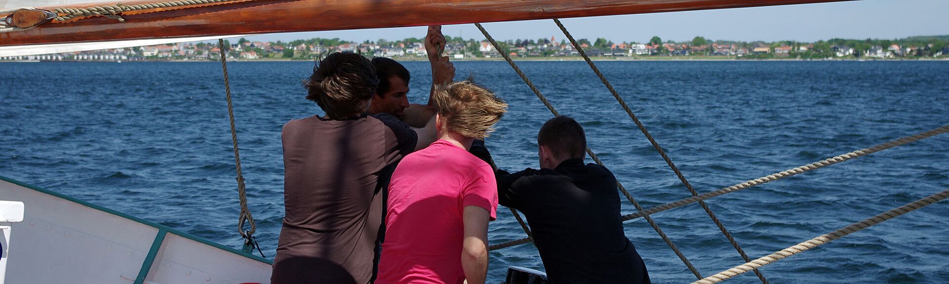 Vier Menschen mit vom Wind zerzausten Haar ziehen auf einem Segelschiff gemeinsam ein Seil an. In der Ferne ist eine Uferlinie mit kleinen Häusern zwischen grünen Bäumen zu sehen.