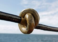 In der schwarzen Strebe der Reling ist ein Knoten eingeschmiedet und in Gold lackiert. Im Hintergrund ist verschwommen Meer, Horizont und Himmel zu sehen.