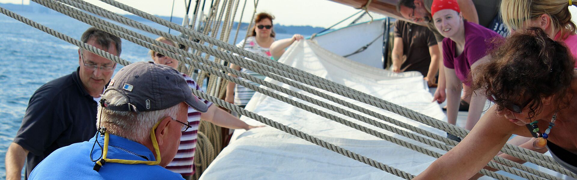 Eine Gruppe von Menschen auf einem Segelschiff faltet an einem sonnigen Tag gemeinsam ein großes, weißes Segel an Deck.