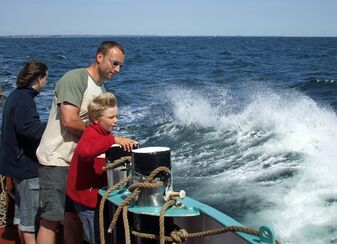 Familienreise auf dem Segelschiff Banjaard: zwei Erwachsene und ein Kind stehen an den Pollern auf dem Vordeck und schauen auf das blaue Wasser, das weißschäumend als Gischt aufspritzt