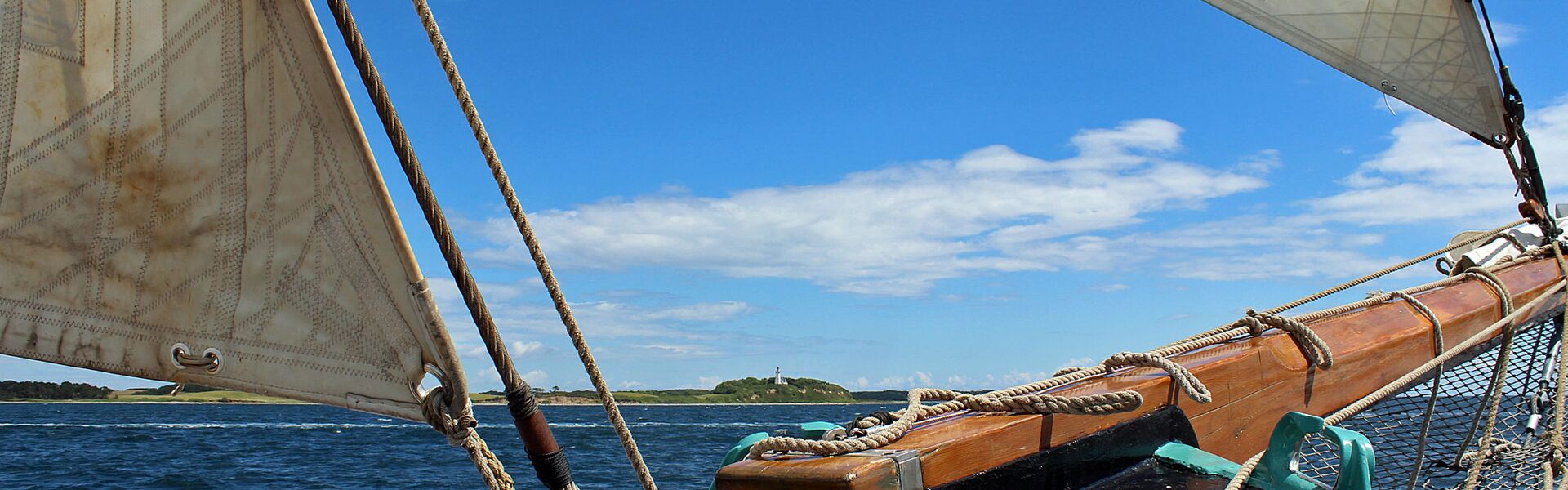 Der hölzerne Klüverbaum des Segelschiffs Banjaard und die unteren Spitzen zweier gesetzter Segel bildet den Rahmen für einen Blick auf die Küste der Insel Samsø mit einem Leuchtturm auf einer Anhöhe.