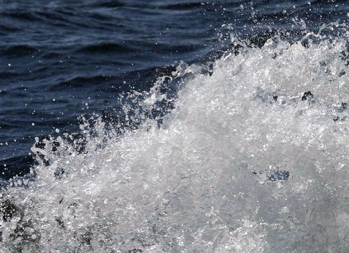 Vor dunkelblauem Wasser spritzt eine Welle in unzählige weiße Tropfen auseinander