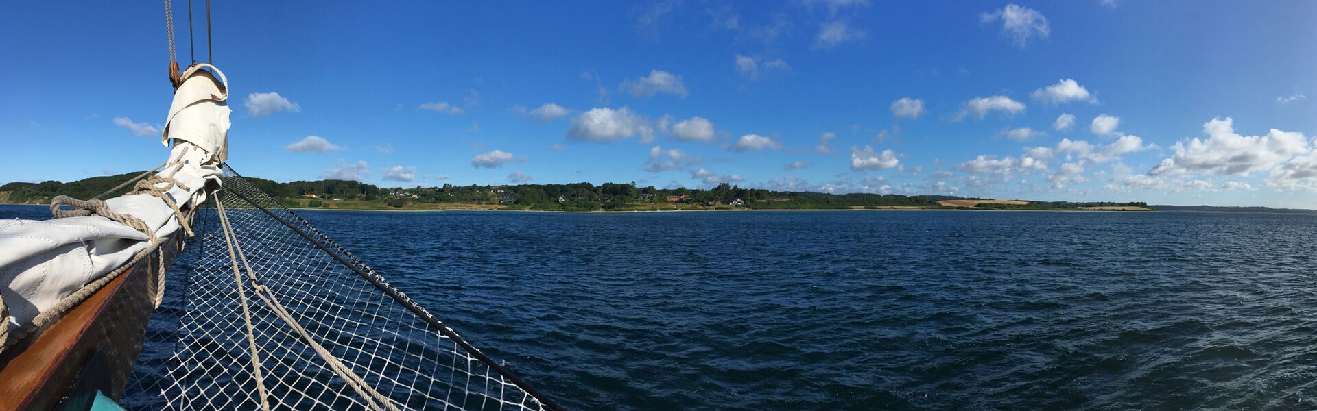 Blick vom Bug des Segelschiffs auf die Küste nahe Ebeltoft in Dänemark, über das stahlblaue Wasser unter einem blauen Himmel mit Tupfenwolken.