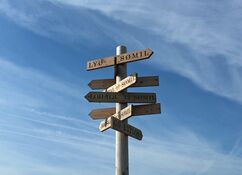 Vor einem blauen Himmel mit weißen Schleierwolken steht ein hölzener Pfahl mit Wegweisern in verschiedene Richtungen, die auf Häfen in der Umgebung zielen und die Entfernung in Seemeilen angeben.