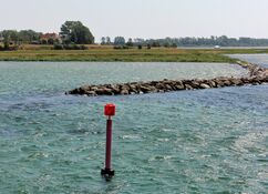 Eine schlanke rote Fahrwasserboje im türkisfarbenen Wasser vor einem Damm aus Natursteinen markiert die Einfahrt zu einem Fjord im Smålandsfahrwasser..