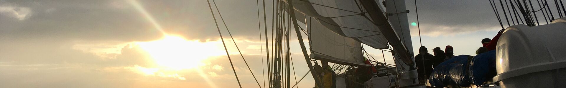 Das Segelschiff Banjaard segelt an einem Morgen im Oktober in den Sonnenaufgang - gegen das Licht sind die Umrisse meherer Personen zu erkennen, die im Bug stehen. Das Licht ist bereits herbstlich.