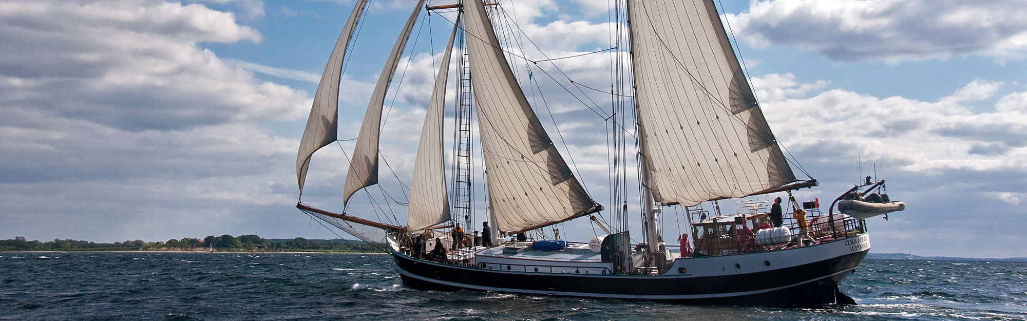Das Segelschiff Banjaard segelt vor der Küste der Kieler Förde unter einem leicht bewölkten Himmel. Im Gegenlicht sind Details wie Nähte und Verstärkungen der weißen Segel betont. An Deck bedienen Menschen konzentriert die Segel und Taue.